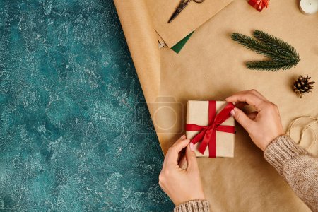 Foto de Vista recortada de la mujer atando lazo de cinta roja en caja de regalo cerca de la decoración de pino sobre fondo texturizado azul - Imagen libre de derechos