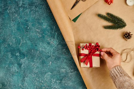 cropped view of man decorating gift box with holly berries on craft paper and blue textured backdrop