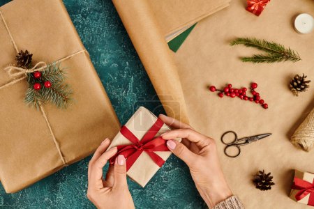abgeschnittene Ansicht einer Frau, die rote Schleife auf Geschenkbox bindet, während sie Weihnachtsgeschenk in der Nähe von Dekorationsartikeln macht