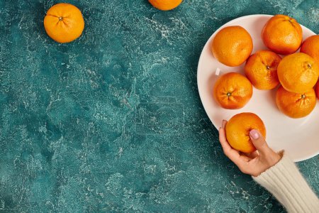 cropped view of woman holding ripe juicy tangerine on blue textured background, Christmas concept