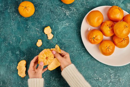 partial view of woman peeling fresh ripe mandarin on blue textured surface, Christmas concept