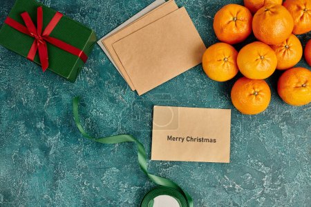 envelope with Merry Christmas lettering near tangerines and gift box with ribbon on blue backdrop