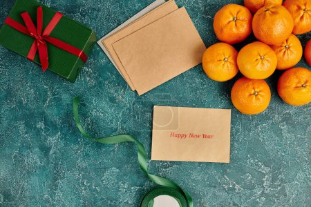 sobre con feliz año nuevo cerca de saludo cerca de mandarines y caja de regalo con cinta sobre fondo azul