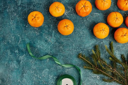frische Mandarinen neben Tannenzweig und dekorativem Band auf blau strukturiertem Hintergrund, Weihnachten