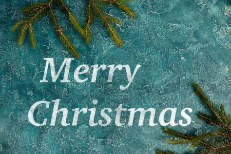 Merry Christmas greeting on blue rustic surface near green pine branches, festive background