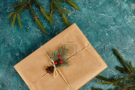 caja de regalo en papel artesanal decorado con bayas de acebo cerca de ramas de pino sobre fondo azul, Navidad