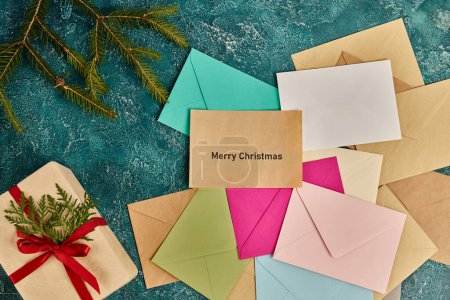 present near pine branches and colorful envelopes on blue rustic backdrop, Merry Christmas lettering