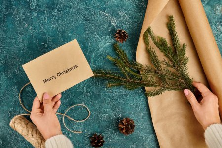 abgeschnittene Ansicht einer Frau mit frohem Weihnachtskuvert und Kiefernzweig in der Nähe von Bastelpapier und Bindfäden, diy