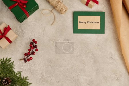 Merry Christmas greeting card near present boxes and juniper branches with holly berries on grey
