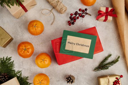 Tarjeta de felicitación con letras de Feliz Navidad cerca de cajas de regalo y decoración festiva sobre fondo texturizado