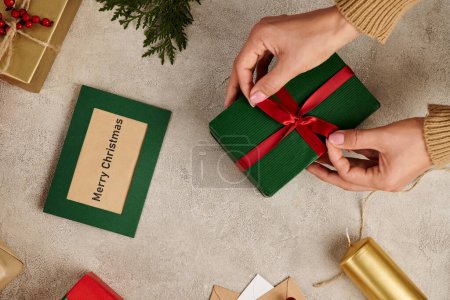 abgeschnittene Ansicht einer Frau, die rote Schleife auf Geschenkbox in der Nähe von Merry Christmas Grußkarte und festlichem Dekor bindet