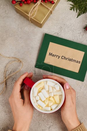partial view of woman holding hot chocolate with marshmallow near Merry Christmas greeting card