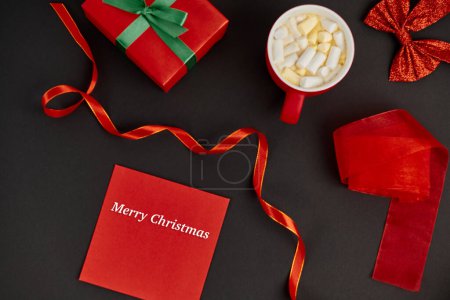 red envelope with Merry Christmas lettering near box and hot chocolate with marshmallows on black