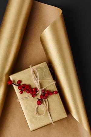 golden gift box with red holly berries on shiny wrapping paper and black backdrop, Christmas decor