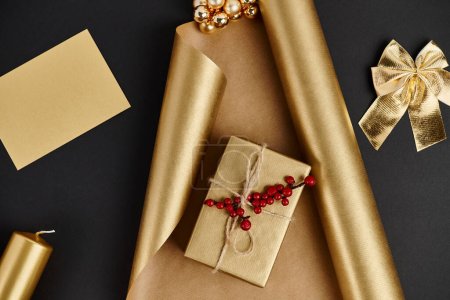 golden Christmas decor, gift box with holly berries on wrapping paper near candle and bow on black