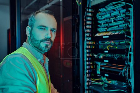 attractive pensive man with beard in safety vest inspecting data center and looking at camera
