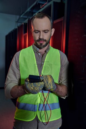 attractive bearded man in safety vest and gloves looking at detector in his hands, data center