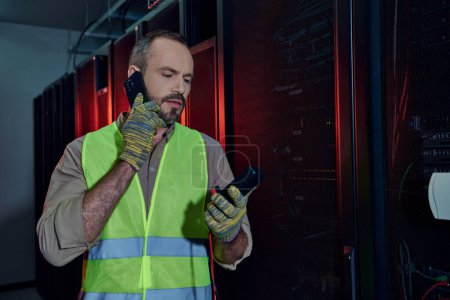 handsome technician with beard in safety vest and gloves holding detector and talking by phone