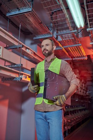handsome concentrated man in safety vest and gloves working hard and looking away, data center