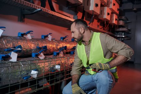good looking pensive technician in safety gloves and vest squatting while working in data center