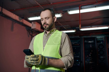 Téléchargez les photos : Beau technicien barbu en gilet de sécurité et gants regardant son smartphone pendant le travail - en image libre de droit