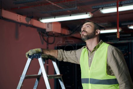 handsome pensive technician in safety vest standing next to step ladder and looking up, data center