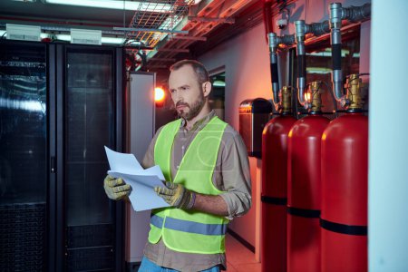 good looking pensive specialist in safety clothes holding papers and looking away, data center
