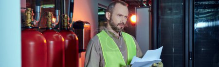 concentrated bearded man in safety vest holding papers and looking away, data center, banner