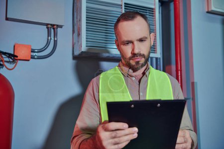attractive pensive professional in safety vest looking at clipboard while working in data center
