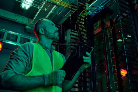 handsome pensive specialist in safety clothes holding clipboard while working hard in data center