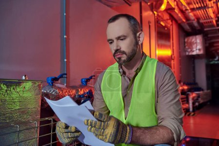 handsome concentrated technician in safety clothes looking attentively at paperwork, data center