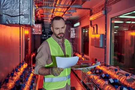concentrated hardworking man in safety clothes looking attentively at his paperwork, data center