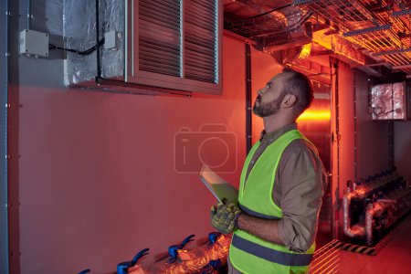 Photo for Attractive concentrated technician in safety gloves and vest holding papers and looking up, data - Royalty Free Image