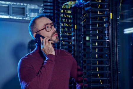 attractive focused specialist in turtleneck with glasses and beard talking by phone, data center