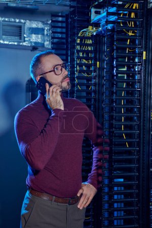 good looking pensive man in turtleneck with glasses with arm on hip talking by phone, data center