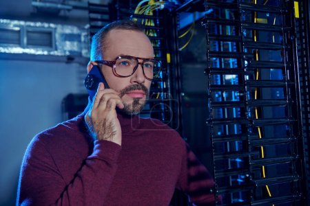 handsome concentrated specialist with glasses and beard talking by phone on work, data center