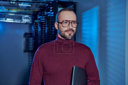 alegre centro de datos guapo especialista en cuello alto con gafas que sostienen el ordenador portátil y mirando hacia otro lado