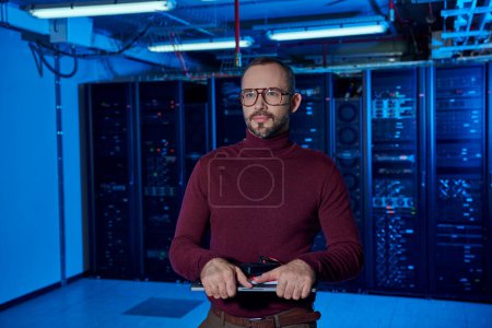 jolly concentrated data center specialist with glasses and beard looking away and holding laptop
