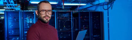 focused jolly specialist in turtleneck holding laptop and looking at camera, data center, banner