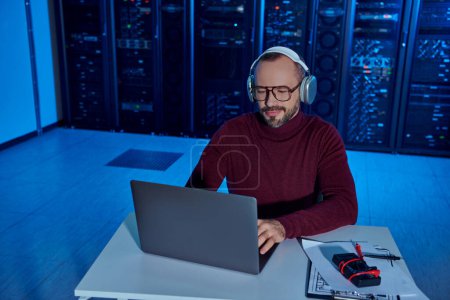 Photo for Joyful concentrated specialist with headphones and glasses sitting and working on his laptop - Royalty Free Image