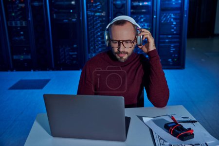 Téléchargez les photos : Spécialiste concentré attrayant avec barbe et écouteurs travaillant dur sur son ordinateur portable, centre de données - en image libre de droit