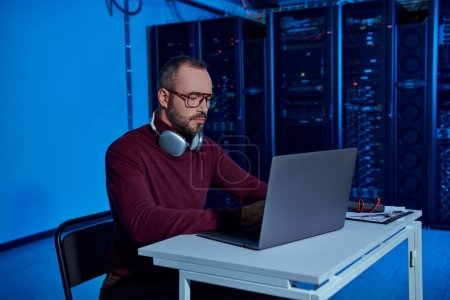 Photo for Handsome concentrated data center specialist sitting at table with headphones and working on laptop - Royalty Free Image