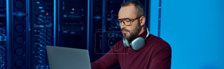 Photo for Focused data center specialist sitting at table with headphones and working on laptop, banner - Royalty Free Image