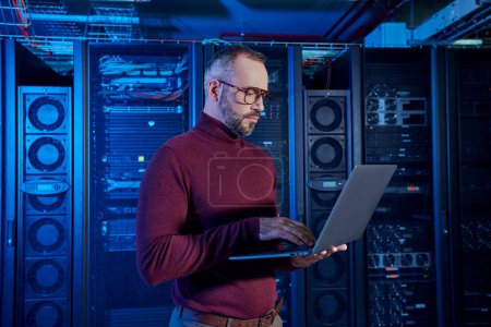 attractive bearded specialist in turtleneck working attentively at his laptop, data center