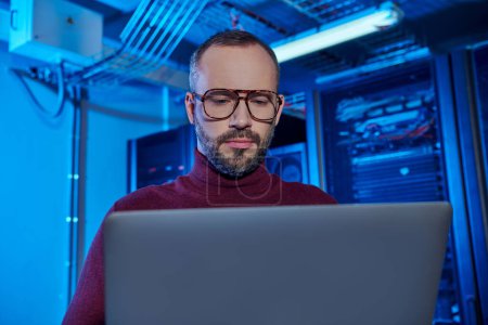 handsome professional with beard and glasses in turtleneck working at his laptop, data center