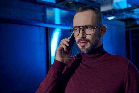 handsome data center specialist with beard and glasses in turtleneck talking by phone at work