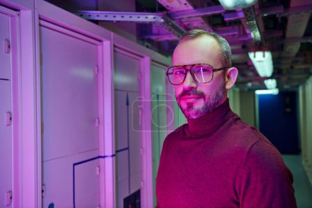 cheerful dedicated specialist in cozy turtleneck with glasses smiling at camera, data center