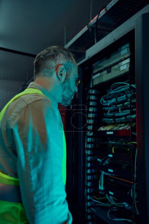 good looking concentrated technician with beard in safety vest working hard in data center