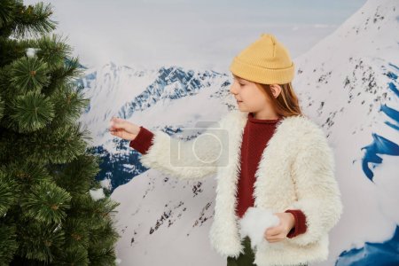 joyful pretty girl in beanie hat and warm attire putting snow on fir tree, fashion concept
