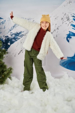 vertikale Aufnahme eines kleinen fröhlichen Mädchens im warmen Winteroutfit mit Beanie-Hut, das mit den Händen zur Seite posiert